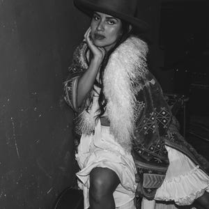 May include: A woman wearing a black hat with a wide brim, a white fur collar, and a long, patterned dress. The dress has a white ruffle at the bottom and a black and white geometric pattern.