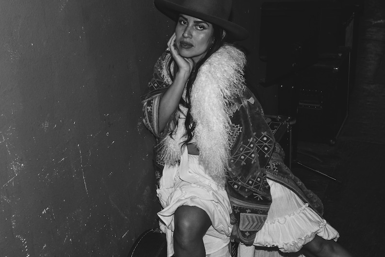 May include: A woman wearing a black hat with a wide brim, a white fur collar, and a long, patterned dress. The dress has a white ruffle at the bottom and a black and white geometric pattern.