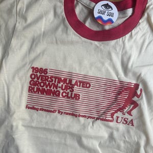 K&ouml;nnte beinhalten: Ein cremefarbenes T-Shirt mit rotem Rundhalsausschnitt und rotem Grafikdruck. Der Text lautet 1986 OVERSTIMULATED GROWN-UPS RUNNING CLUB, Feeling stressed? Try running from your problems! USA, neben der Silhouette einer laufenden Person.