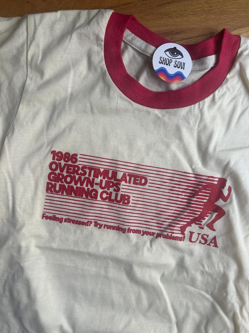 K&ouml;nnte beinhalten: Ein cremefarbenes T-Shirt mit rotem Rundhalsausschnitt und rotem Grafikdruck. Der Text lautet 1986 OVERSTIMULATED GROWN-UPS RUNNING CLUB, Feeling stressed? Try running from your problems! USA, neben der Silhouette einer laufenden Person.