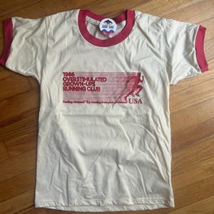 K&ouml;nnte beinhalten: Ein cremefarbenes Ringer-T-Shirt mit rotem Besatz an Kragen und &Auml;rmeln. Die Vorderseite zeigt eine rote Grafik mit dem Text 1986 OVERSTIMULATED GROWN-UPS RUNNING CLUB, die Silhouette eines L&auml;ufers und den Text Feeling stressed? Try running from your problems! USA.
