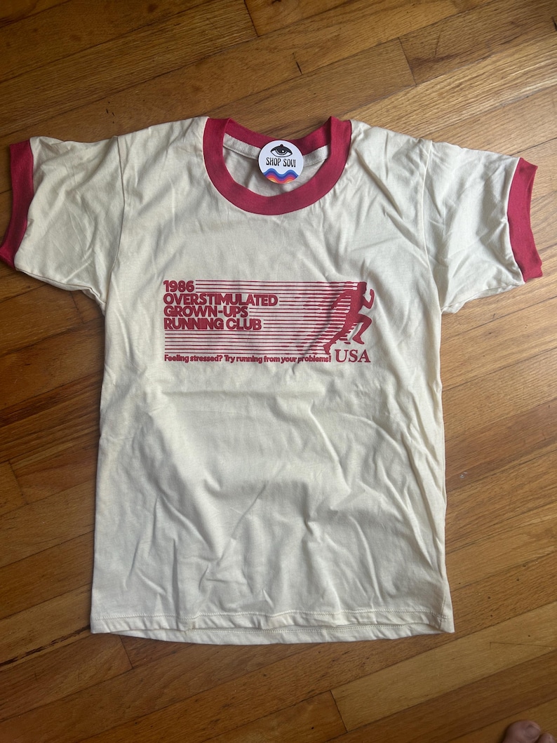 K&ouml;nnte beinhalten: Ein cremefarbenes Ringer-T-Shirt mit rotem Besatz an Kragen und &Auml;rmeln. Die Vorderseite zeigt eine rote Grafik mit dem Text 1986 OVERSTIMULATED GROWN-UPS RUNNING CLUB, die Silhouette eines L&auml;ufers und den Text Feeling stressed? Try running from your problems! USA.