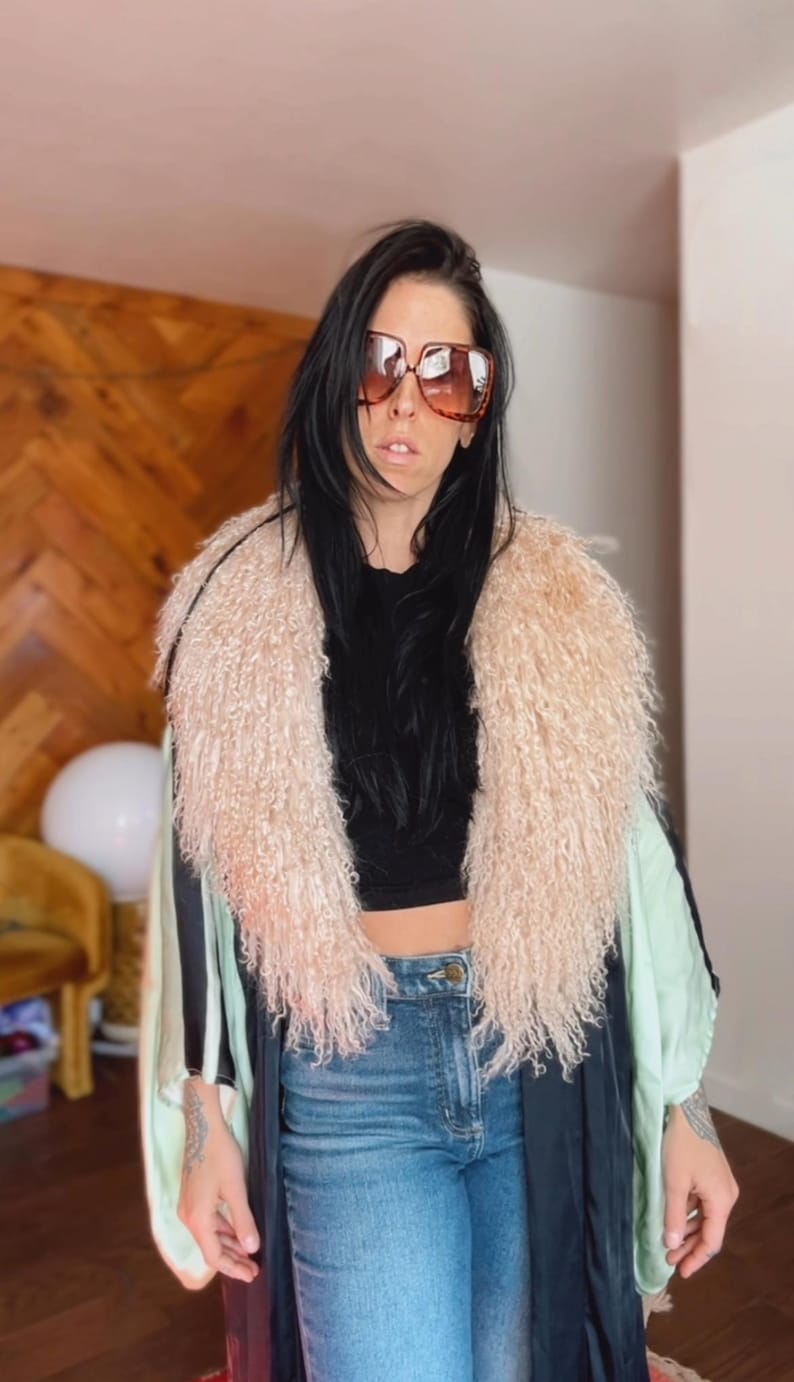 May include: A woman wearing a black and green kimono with a fluffy white collar. She is wearing blue jeans and brown sunglasses.