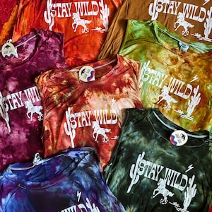 May include: A collection of six tie-dye tank tops with the phrase "Stay Wild" printed in white on each. The tank tops are in various colors, including orange, red, purple, green, and blue.