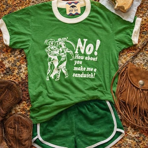 May include: A green ringer t-shirt with white trim and a white graphic print showing two figures and the text "SLAP! No! How about you make me a sandwich!" paired with green athletic shorts featuring white trim along the curved leg openings.
