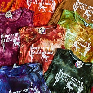 May include: A collection of tie-dye tank tops with the phrase "Stay Wild" printed in white on each. The tops are in various colors, including orange, green, purple, and brown. Each top features a cactus and a horse design.
