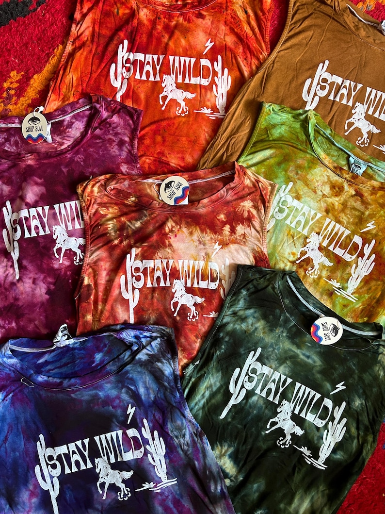 May include: A collection of tie-dye tank tops with the phrase "Stay Wild" printed in white on each. The tops are in various colors, including orange, green, purple, and brown. Each top features a cactus and a horse design.