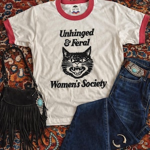 May include: A white ringer t-shirt with pink trim featuring a black graphic of a snarling cat and the text "Unhinged & Feral Women's Society". The outfit includes dark blue denim flare jeans, a black fringed handbag, and turquoise jewelry.