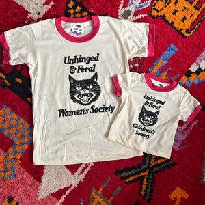May include: Two off-white ringer t-shirts with pink trim. The larger shirt features a black cat graphic with the text "Unhinged & Feral Women's Society". The smaller shirt features a similar cat graphic with the text "Unhinged & Feral Children's Society".
