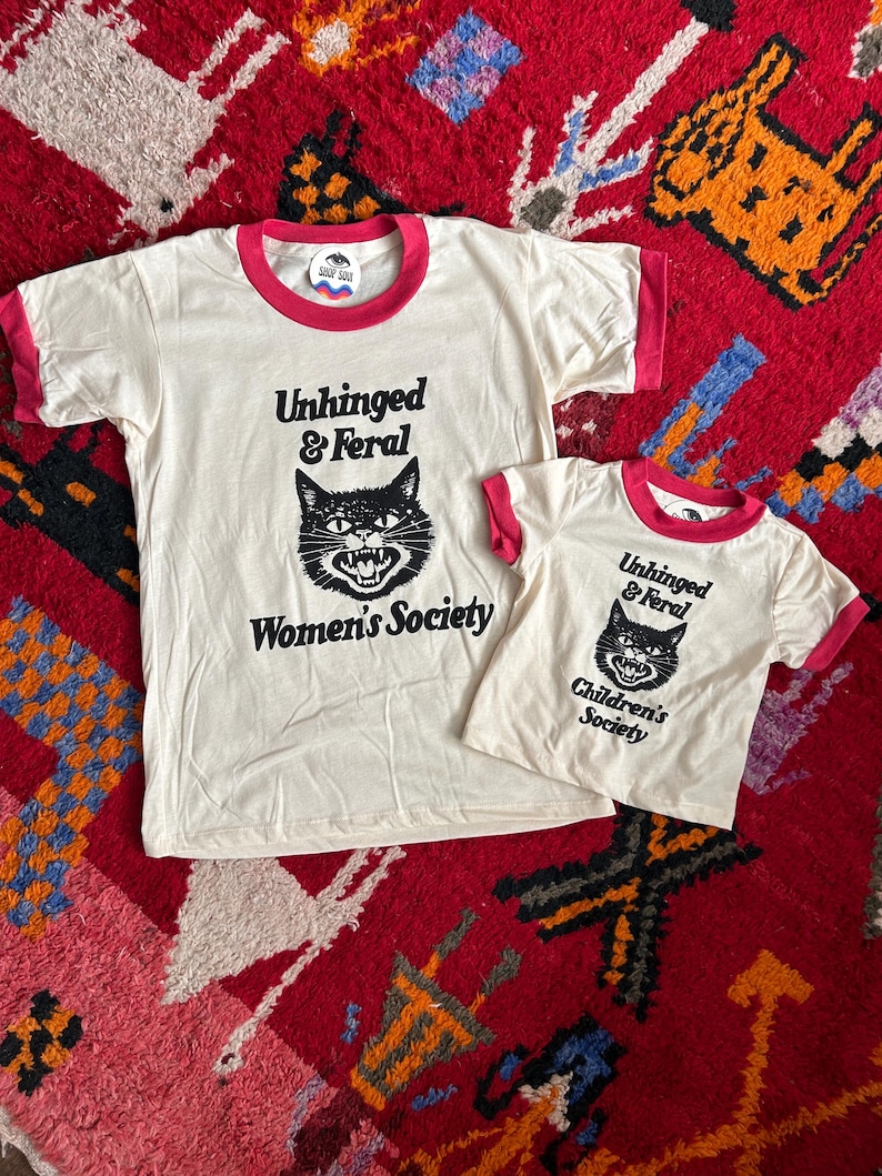 May include: Two off-white ringer t-shirts with pink trim. The larger shirt features a black cat graphic with the text "Unhinged & Feral Women's Society". The smaller shirt features a similar cat graphic with the text "Unhinged & Feral Children's Society".