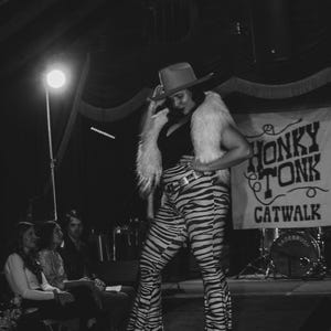 May include: A woman wearing a black and white zebra print pantsuit with a white fur vest and a cowboy hat. She is standing on a stage with a sign that says "Honky Tonk Catwalk".