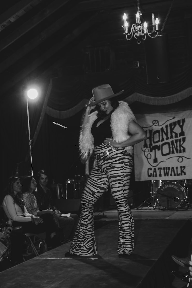 May include: A woman wearing a black and white zebra print pantsuit with a white fur vest and a cowboy hat. She is standing on a stage with a sign that says "Honky Tonk Catwalk".