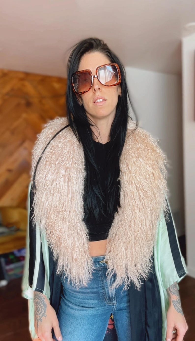 May include: A woman wearing a black top, blue jeans, and a long, light green and black kimono with a fluffy, light pink collar. She is wearing large, brown, square-shaped sunglasses.