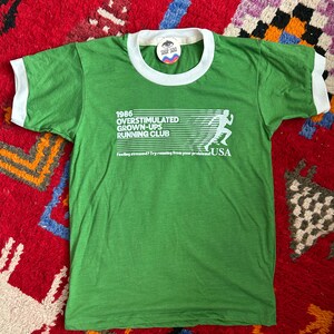 K&ouml;nnte beinhalten: Ein gr&uuml;nes Ringer-T-Shirt mit wei&szlig;em Besatz an Kragen und &Auml;rmeln. Auf der Vorderseite befindet sich der wei&szlig;e Schriftzug 1986 OVERSTIMULATED GROWN-UPS RUNNING CLUB, eine Grafik einer laufenden Person und der Text Feeling stressed? Try running from your problems! USA.