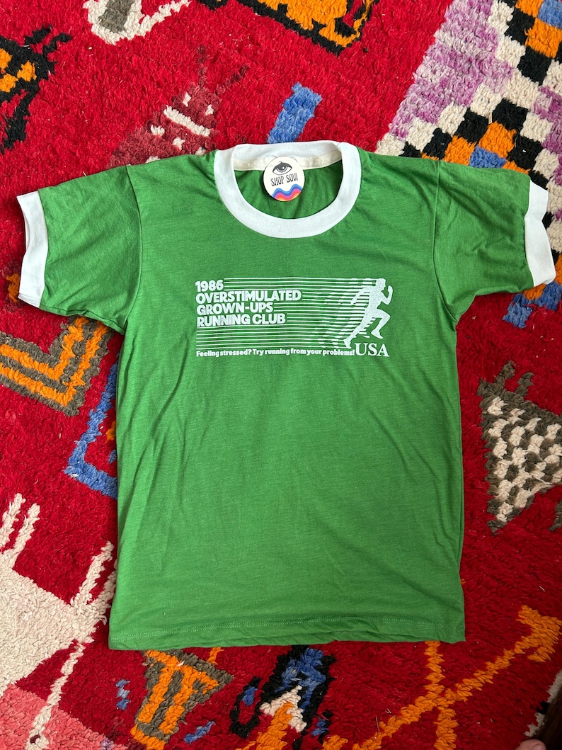 K&ouml;nnte beinhalten: Ein gr&uuml;nes Ringer-T-Shirt mit wei&szlig;em Besatz an Kragen und &Auml;rmeln. Auf der Vorderseite befindet sich der wei&szlig;e Schriftzug 1986 OVERSTIMULATED GROWN-UPS RUNNING CLUB, eine Grafik einer laufenden Person und der Text Feeling stressed? Try running from your problems! USA.
