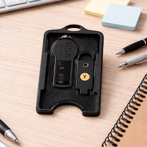May include: A black 3D-printed plastic holder containing an RSA security token with a digital display and a YubiKey with a gold circular button featuring a lowercase y. The holder has a rectangular frame with a top hole for a lanyard or keychain.