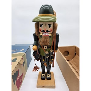 Puede incluir: Figura de cascanueces de madera de un cazador con un atuendo verde y negro, que lleva un sombrero con una pluma. La figura tiene barba y bigote de piel sintética, y sostiene unos prismáticos y una bolsa. Se encuentra sobre una base de madera.