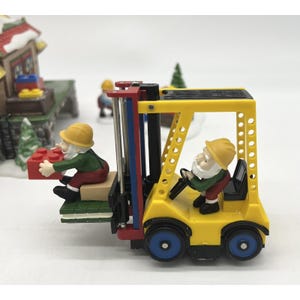 Könnte beinhalten: Eine Miniatur-Weihnachtsdorf-Szene mit einem gelben Gabelstapler mit rotem und schwarzem Rahmen. Zwei Zwerge bedienen den Gabelstapler, einer lädt einen roten Block. Die Szene umfasst ein Gebäude und einen kleinen Weihnachtsbaum.