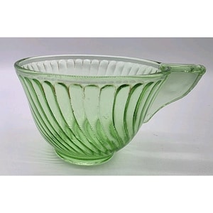 Può includere: Una tazza da tè vintage in vetro verde con manico. La tazza presenta un motivo a spirale all'esterno e una forma svasata. Il vetro è di colore verde chiaro e la tazza sembra essere in buone condizioni.