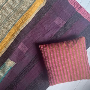 Peut inclure: Une courtepointe patchwork violette et dorée avec un coussin rayé rose et doré sur le dessus. La courtepointe est faite de différents tissus et a un aspect rustique et vintage.