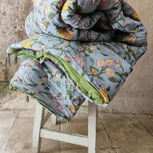 Könnte beinhalten: Eine hellblaue Blumensteppdecke mit gelben, rosa und weißen Blumen. Die Steppdecke ist gefaltet und auf einem kleinen, weißen Holzschemel platziert. Die Steppdecke hat einen grünen Rand und Quasten.