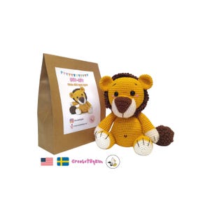 Op de afbeelding: Een handgemaakt gehaakt leeuwenspeelgoed met een bruine manen, staart en neus en witte poten. De leeuw is geel en bruin. Links van de leeuw staat een bruine papieren zak met een DIY-kit label.