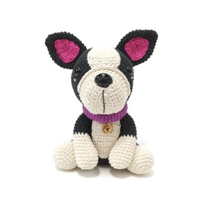 May include: A handmade crochet Boston Terrier dog with black, white, and pink details. The dog has a purple collar with a gold charm. The dog is sitting upright with its paws in front. The background is white.