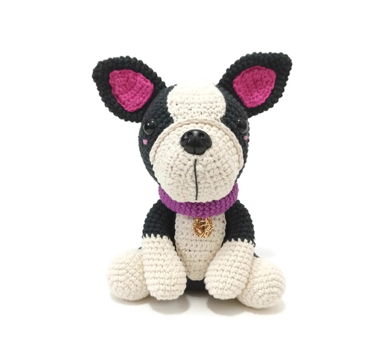May include: A handmade crochet Boston Terrier dog with black, white, and pink details. The dog has a purple collar with a gold charm. The dog is sitting upright with its paws in front. The background is white.
