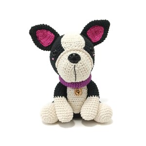 May include: A crocheted Boston Terrier dog with black, white, and pink details. The dog has a purple collar with a gold charm. The dog is sitting upright with its paws in front.