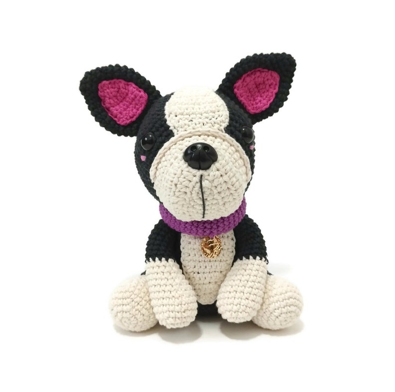 May include: A crocheted Boston Terrier dog with black, white, and pink details. The dog has a purple collar with a gold charm. The dog is sitting upright with its paws in front.