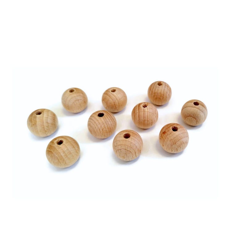 May include: Ten natural wood beads, each with a hole in the center, are scattered on a white surface. The beads are round and smooth, and they are all the same size.