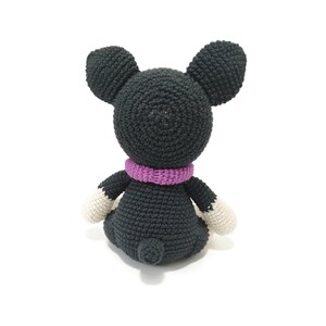 May include: A crocheted mouse toy, seen from the rear, made from dark grey yarn. It features large, rounded ears, a purple scarf, and white paws. The toy is sitting on a white surface.