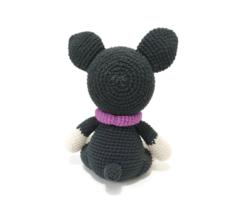 May include: A crocheted mouse toy, seen from the rear, made from dark grey yarn. It features large, rounded ears, a purple scarf, and white paws. The toy is sitting on a white surface.