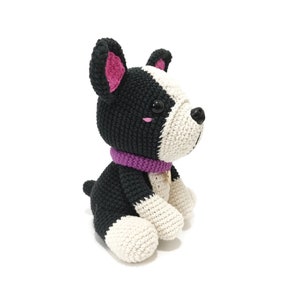 May include: A crocheted dog toy in black and white, with a purple collar. The dog has black ears with pink inner detail, and a white face. The dog is sitting with its paws forward, and has a small gold tag on its collar.