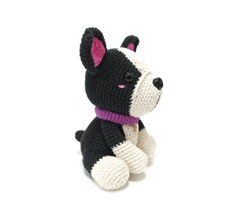 May include: A crocheted dog toy in black and white, with a purple collar. The dog has black ears with pink inner detail, and a white face. The dog is sitting with its paws forward, and has a small gold tag on its collar.