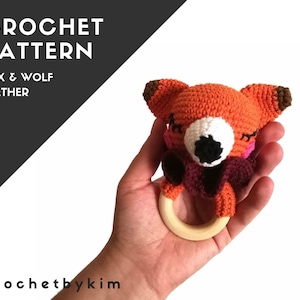 May include: Crochet pattern for a fox or wolf teether. The teether is orange, brown, white, and pink. It has a wooden ring and is held in a hand.