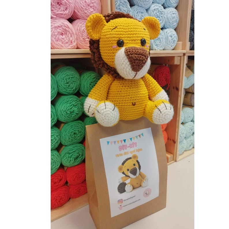 Op de afbeelding: Een handgemaakte gele gehaakte leeuw met een bruine manen en witte accenten zit bovenop een bruine papieren zak. De zak heeft een label met de tekst "DIY-KIT" en een kleine leeuwenillustratie. Op de achtergrond zijn planken met kleurrijk garen te zien.