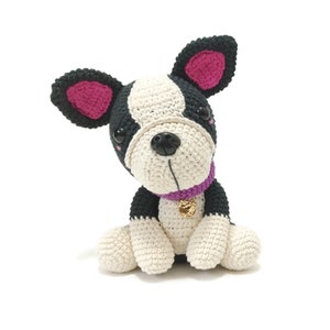 May include: A crocheted dog toy in black, white, and pink. The dog has a black nose, black eyes, and a purple collar with a gold charm. The ears are black with pink insides. The toy is sitting on a white surface.