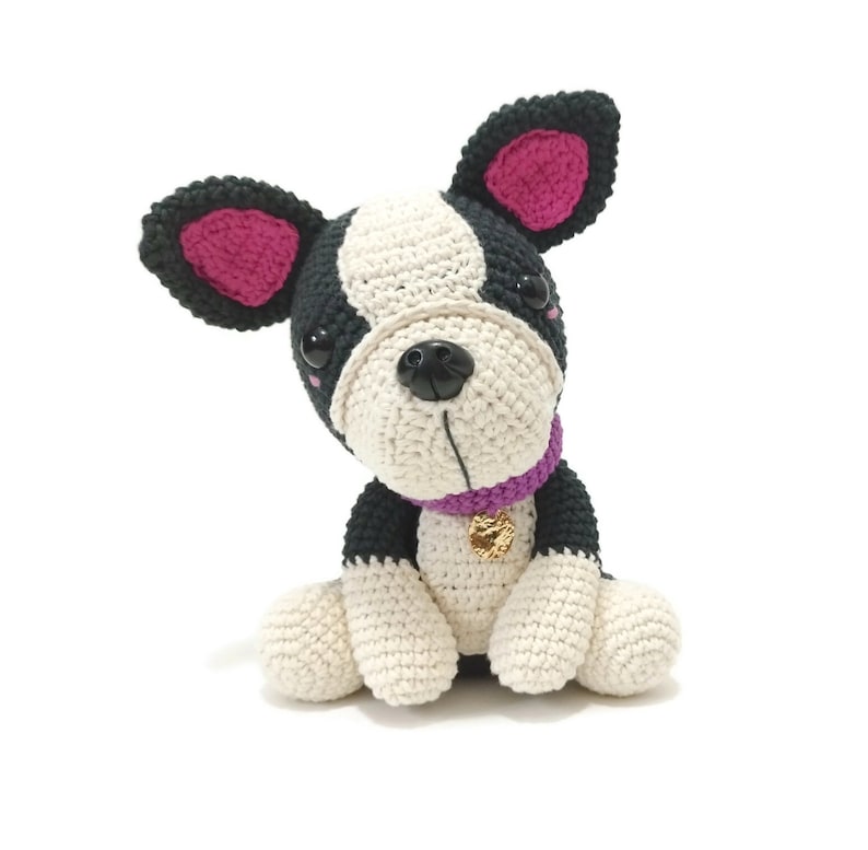 May include: A crocheted dog toy in black, white, and pink. The dog has a black nose, black eyes, and a purple collar with a gold charm. The ears are black with pink insides. The toy is sitting on a white surface.