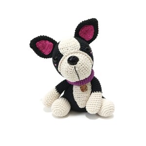 May include: A crocheted dog toy in black, white, and beige. The dog has a purple collar with a gold charm and pink inner ears. The dog is sitting with its head tilted, showing its black button eyes and black nose.