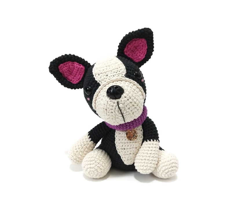 May include: A crocheted dog toy in black, white, and beige. The dog has a purple collar with a gold charm and pink inner ears. The dog is sitting with its head tilted, showing its black button eyes and black nose.