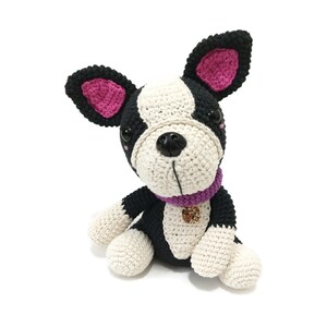 May include: A handmade crochet Boston Terrier dog with black, white, and pink details. The dog has a purple collar with a gold charm. The dog is sitting on a white surface.