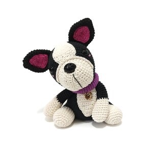May include: A crocheted dog toy in black, white, and cream. The dog has a purple collar with a gold charm. The ears are lined with pink. The dog is sitting and looking to the side.