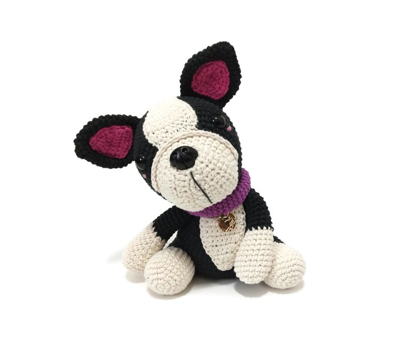 May include: A crocheted dog toy in black, white, and cream. The dog has a purple collar with a gold charm. The ears are lined with pink. The dog is sitting and looking to the side.