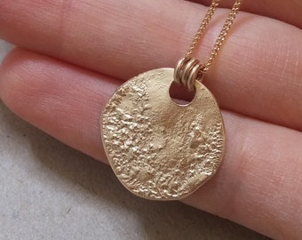 Könnte beinhalten: Eine goldene Halskette mit einem strukturierten runden Anhänger. Der Anhänger hat ein kleines Loch in der Mitte.