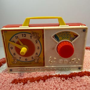 Puede incluir: Reloj de juguete Fisher-Price vintage, rojo y amarillo. La esfera del reloj tiene una manecilla amarilla e ilustraciones de ratones. El lado derecho tiene un dial con el texto "Hickory Dickory Dock". El juguete tiene un asa amarilla. El texto "FISHER-PRICE TOYS" es visible.