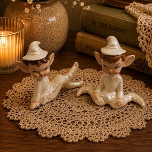 Peut inclure: Deux figurines d'elfes en céramique avec une finition blanche irisée, des chapeaux pointus et des cheveux bruns. Un elfe est allongé sur le côté, tandis que l'autre est assis avec les jambes étendues. Les deux présentent des détails peints.