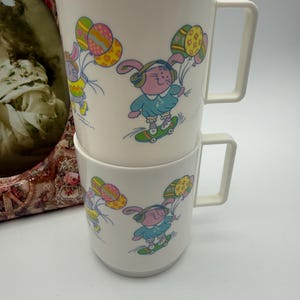 Könnte beinhalten: Zwei weiße Tassen, die übereinander gestapelt sind. Jede Tasse zeigt einen Cartoon-Hasen, der Skateboard fährt und Ostereier-Ballons hält. Der Hase trägt Kopfhörer und ein blaues Outfit. Die Tassen haben eckige Griffe.