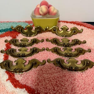 Peut inclure: Six poignées de tiroir antiques en laiton avec des motifs ornés sont présentées sur un tapis rose et blanc. Un récipient jaune rempli de pêches est en arrière-plan. Pour meubles.