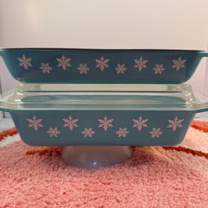 May include: Two teal rectangular casserole dishes with snowflake designs. The top dish has a black rim and is stacked on top of a similar dish with a clear glass lid. The dishes are on a white pedestal.
